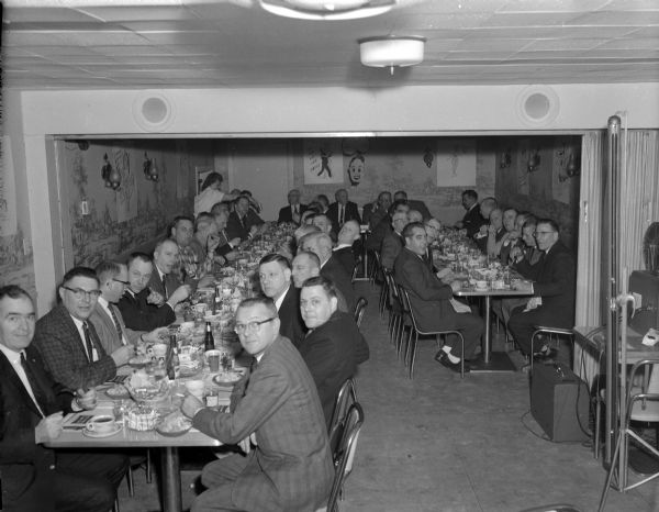 Elevated view of a large group of men sitting down at a fish banquet. The men are sitting at long tables spanning two rooms. On the far right is a table with a film reel in a film projector, and a folded screen. probably a dinner meeting of the "Ice Chippers".