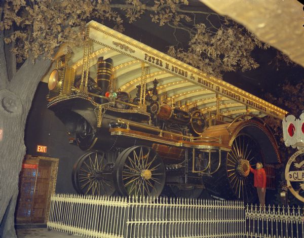 A woman stands next to a locomotive on display behind a fence at House on the Rock. This image was included in the attraction's brochure. The sign on the train reads: "Peter H. Burno. Mighty in Strength & Endurance." There is a large, fake tree on the left.