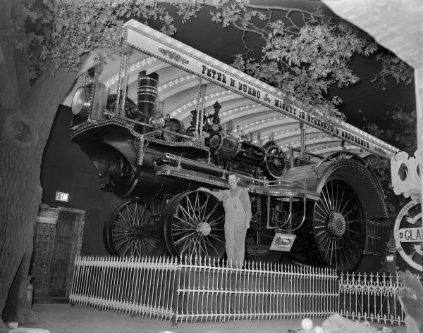 Peter Burno posing with the steam locomotive he built which is on display behind a fence at House on the Rock. The sign above the train reads: "Peter H. Burno. Mighty in Strength & Endurance." There is a large, fake tree on the left.