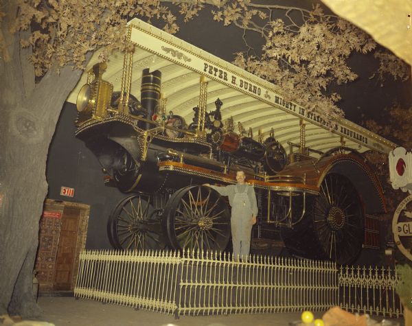 A man posing with a locomotive on display behind a fence at House on the Rock. The sign on the train reads: "Peter H. Burno. Mighty in Strength & Endurance." There is a large, fake tree on the left.