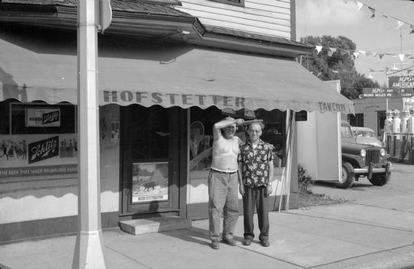 View from street of two men standing on the sidewalk in front of the Hofstetter tavern. The Gisholt sales representative standing on the left is holding a giant comb over the bare head of the man standing next to him. There is a gas station next door on the right.