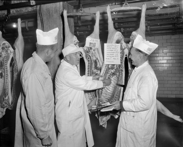 Sid talking to two employees in the Oscar Mayer Company. All three men are wearing long white coats and hats. Sid is standing in the middle next to meat hanging from meat hooks. There are signs attached to each piece of meat.