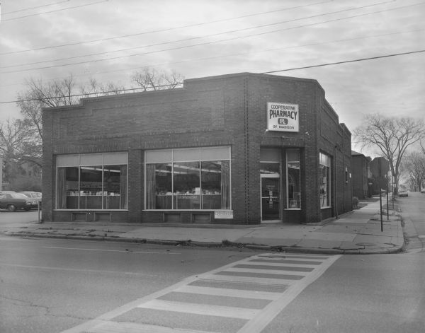 View across street towards the Cooperative Pharmacy of Madison. The pharmacy is in a brick building on a corner. Cars are parked in a lot on the left.
