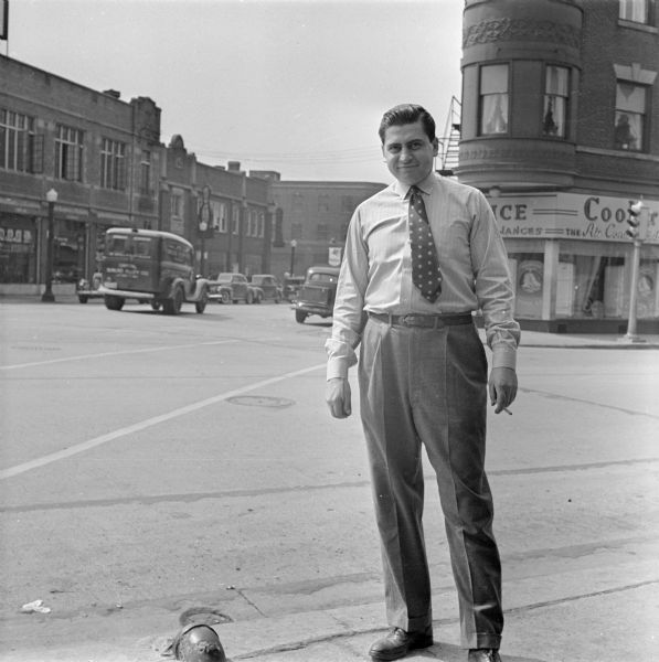 View of a man standing on the sidewalk near State and Gorham streets holding a cigarette in his hand. Behind him is an intersection with traffic and cars parked on both sides of the street near storefronts. 