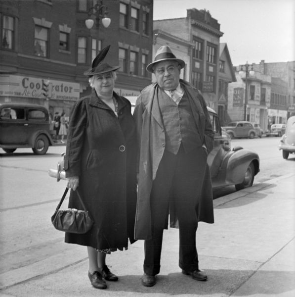 A man and a woman standing and posing together on a sidewalk near State and Gorham Streets. (Oscar Mayer's Coolerator store was located at 401-05 State Street) Behind them is an intersection with traffic and cars parked on both sides of the street near storefronts.  The couple might be Sid Boyum's aunt and uncle Miss Martha Schroeder and George Schroeder, his mother's sister and brother. 