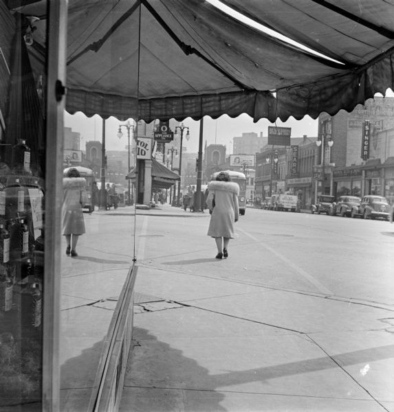 View from under the Badger Liquor awning over a storefront of a woman crossing an intersection at State Street. Across the street on the right are storefronts along the street, with signs including "Singer" and "Sears." Further up the street is the Capitol Theatre tower and the Wisconsin State Capitol.