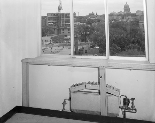 Elevated view (possibly) from a room through the window of the UW-Extension building on Lake Street, looking towards the Wisconsin State Capitol. The Orpheum Theatre sign and the Capitol Theater tower are just above the trees in the foreground. Men are working on the roof of a large building under construction on the left.