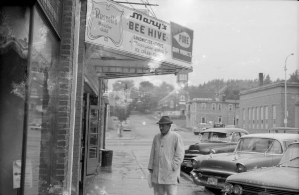Man on the sidewalk in front of Mary's Bee Hive, a lunch counter. Cars are parked at an angle to the curb on the right.