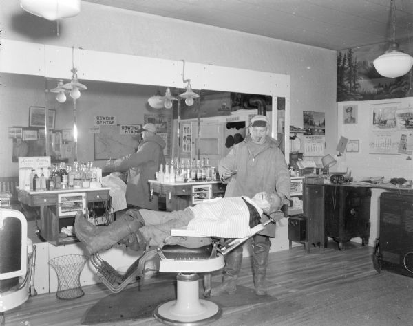 Sid giving a man a shave in Moon's Barber Shop. Sid has a cigar in his mouth. The man sitting in the chair getting the shave is wearing hip waders. A large mirror is on the wall in the background.