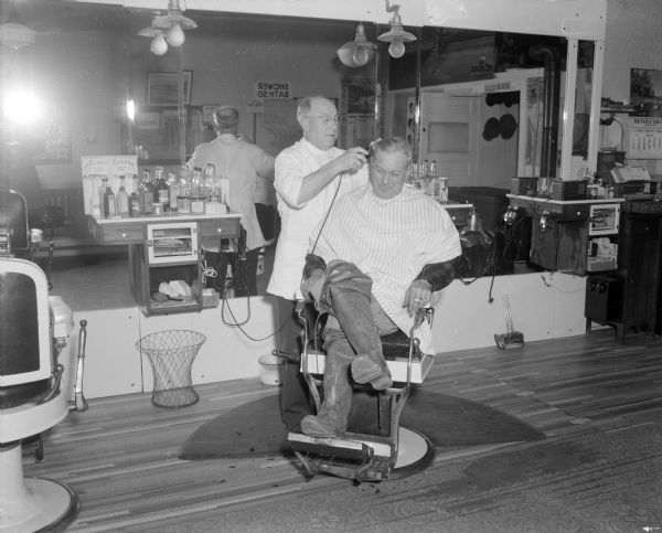 Barber shaving the head of a man in Moon's Barber Shop. The man getting shaved is holding a cigarette and is wearing hip waders rolled down over his knees. A large mirror is on the wall in the background, and on the far left is the reflection of the photographer holding the camera with a flash.