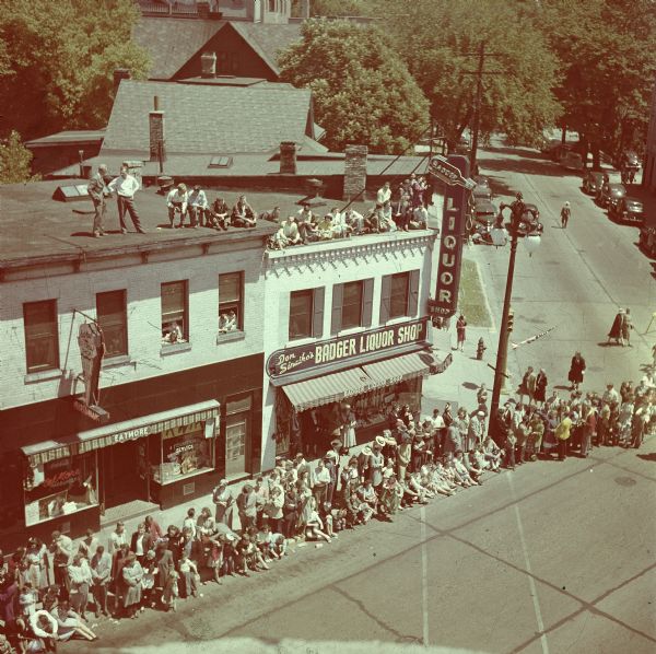Elevated view across the street of the Badger Liquor Shop at the intersection of State and Gorham Streets. People are standing and sitting along the curb in front of the liquor store. A group of people are gathered on the roof of the liquor shop and also the roof above the "Eatmore" storefront. Parked cars, houses and trees are in the background.