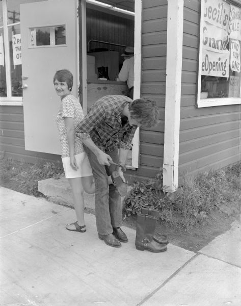 Grand opening of Cecil's Sandals. Judy Pellett, daughter of Cecil Burke and Isabel Burke,founders of Cecil’s Sandals, is wearing shorts and sandals and is standing outside the open door of Cecil's, her right leg extended backward.  Jerry Smotherman, an employee at Cecil's is wearing a plaid shirt and is standing back-to-back with her holding her right sandal between his knees and holding a hammer above the sole. A pair of harness boots is standing on the sidewalk near the man's feet. Sid Boyum is seated inside the shop.