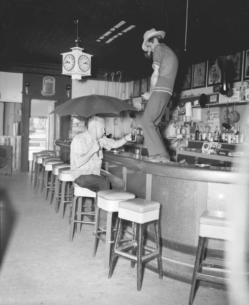 Sid sitting on a stool at the Karabis Bar with a cigar in his mouth. He is holding an umbrella in his right hand and a drink in his left hand, being filled provocatively by  the bartender who stands above Sid on the top of the bar. Letters attached to the ceiling read: "If You Drink To Forget, Please Pay In Advance."