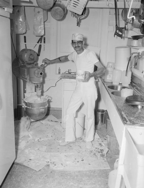 A man wearing a white t-shirt, white pants, and paper hat, is standing and holding a container in his left hand and operating a lever with his right hand for an industrial mixer full of Tom & Jerry Batter in the Karabis Tavern preparation area. His wristwatch is pulled high up on his right forearm. Printed on the container are the words: "George & Tom's Lickin' Good Tom & Jerry Batter."