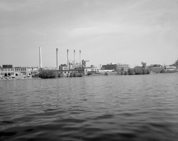 View across water towards the shoreline of the eastern isthmus of Madison, including the MG&E power plant smokestacks. A group of people appear to be fishing in front of The Madison Elks Lodge building on the far right shoreline.
