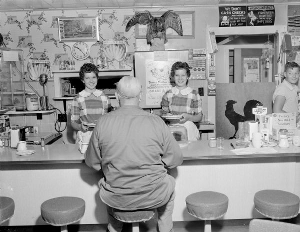View of a man sitting on a stool at the counter of the diner. Behind the counter are standing two uniformed waitresses, perhaps twins, each holding plates of food and smiling. Behind the counter on the right a young man is turning and watching.
