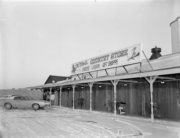View from parking lot of "Del Wood's Country Store, Cheese, Liquor, Gift Shoppe." Three people are standing on the covered porch near the entrance. Agricultural implements are displayed on the porch. Snow is on the ground, and two silo's are behind the store on the far right.