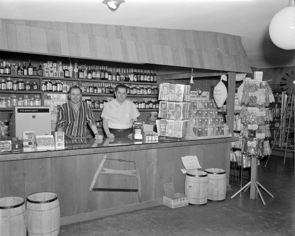 Two men standing behind the counter near the cash register at Del Wood's Country Store. Behind them are shelves of liquor bottles and cigarettes, and on the counter are displays of candy and greeting cards, and a jar of pickles.