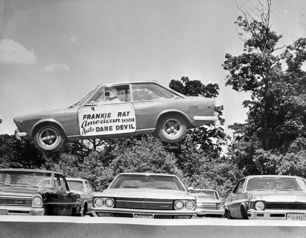 Photographic cutout of a man driving an automobile pasted over another image of parked cars. The automobile appears to be flying over the row of cars. The man is wearing a white helmet that reads: "DAREDEVIL." A sign on the driver's side door reads: "Frankie Ray, American Auto Dare Devil."  