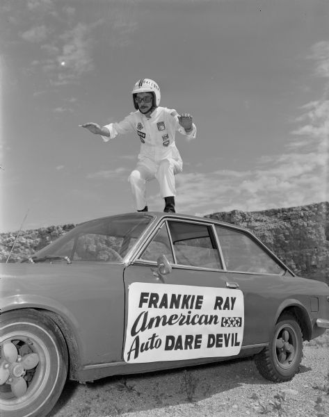 A man is standing on the top of a racing car and posing as if the car is in motion. The man is wearing a white jumpsuit with patches on the chest, eyeglasses, black boots, and a white helmet that reads in part: "AUTO DAREDEVIL." A sign on the driver's side door reads: "Frankie Ray, American Auto Dare Devil." In the background is a rock wall, perhaps taken in a quarry.