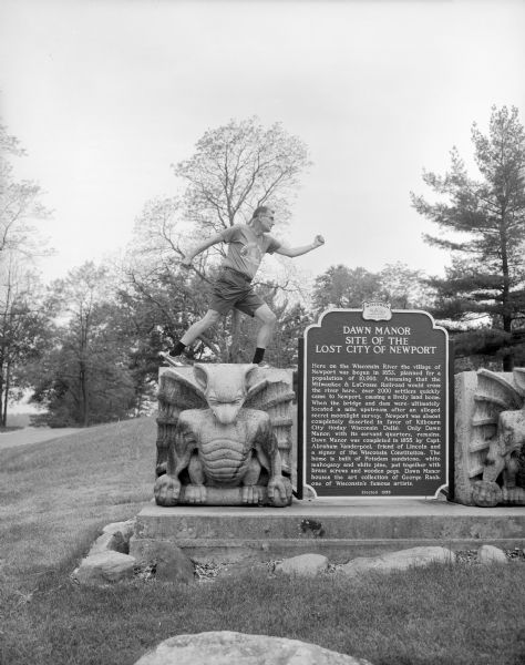 A man is posing in a running profile on top of one of two stone gargoyles that flanks an historical marker for the Dawn Manor in the Wisconsin Dells. Dedicated in 1955, the marker title reads: "Dawn Manor, Site of the Lost City of Newport." The gargoyles in relief represent those purchased and transported from Chicago in 1929 by W.J. Newman, the third owner of the manor. Helen H. Raab purchased the manor in 1942 and chose what she likened to stone bats to stand on either side of the Dawn Manor historical marker, because when she took ownership of the house it was full of bats.
