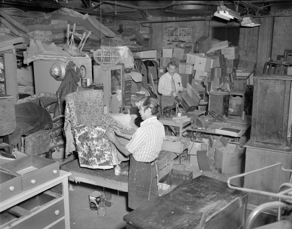Elevated view of two clerks wearing aprons working in the storehouse of the thrift store. One man is reupholstering an old armchair, and the other man is holding an old wooden case. The room is crowded with donated secondhand furniture, clothes and other items. In the back of the room is a stack of boxes of unprocessed donated items. In the center of the room between the two men is a sewing machine and other tools.