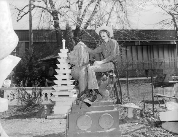 Portrait of Jerry Smotherman, an employee of Cecil's Sandals and Boot Ranch, sitting on a western saddle placed over the back of the Southeast Asian-styled "Guardian Lion" sculpture in Sid's backyard. The man is wearing a hat, blue jeans and a pair of cowboy boots produced by Cecil's Sandals. He is sitting beside the white "Multi-tiered Pagoda." 