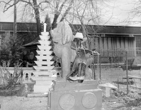 A cowboy hat, a western saddle and a pair of cowboy boots produced by Cecil's Sandals on the Southeast Asian-styled "Guardian Lion" sculpture in Sid's backyard. 