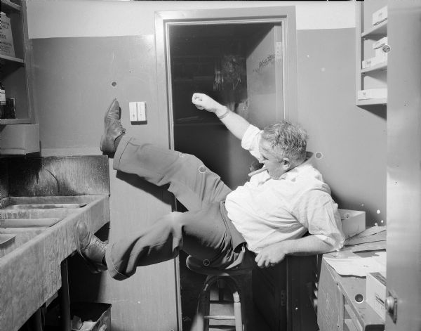 Sid simulating falling in the darkroom with a rather awkward posture. He is sitting on a stool with his legs up in the air, and is pretending to hold something with his right hand.
