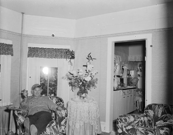 Sid sitting with his feet up in an armchair. He is wearing a hat and is holding a cigar in his right hand. There is a flower arrangement on a table on the right, which is near the door of the kitchen. An older woman is sitting on a chair in the kitchen. The flash of the camera used to take this picture can be seen in the window behind him. 