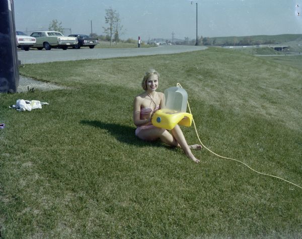 Faye Geise, wearing a bikini, is sitting on a lawn and posing with Aqua Form, a local invention. 