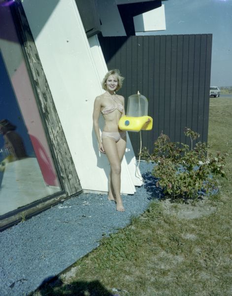 Faye Geise, wearing a bikini, is standing outdoors and posing with Aqua Form, a local invention. She is standing on colored gravel next to a building with a large window.