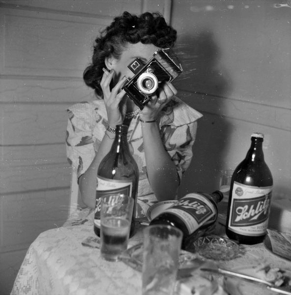 Portrait of a woman sitting on a chair holding a cigarette and holding a camera up to her eye. On the table in front of her are three Schlitz beer bottles, glasses and an ashtray.