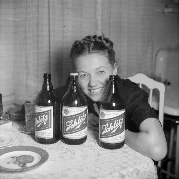 Portrait of a smiling woman, sitting behind three Schlitz beer bottles on a table in front of her. 
