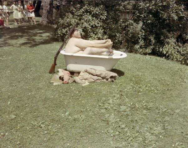 Len Nyberg sitting in a white, enamel metal bathtub set up on a lawn outdoors near the Yahara River. He is washing his feet. A wooden duck is floating in the tub which is full of water, and there is a shotgun placed upright against the end of the tub by his head. His clothes are lying on the grass. In the background, a group of women are watching.                                        