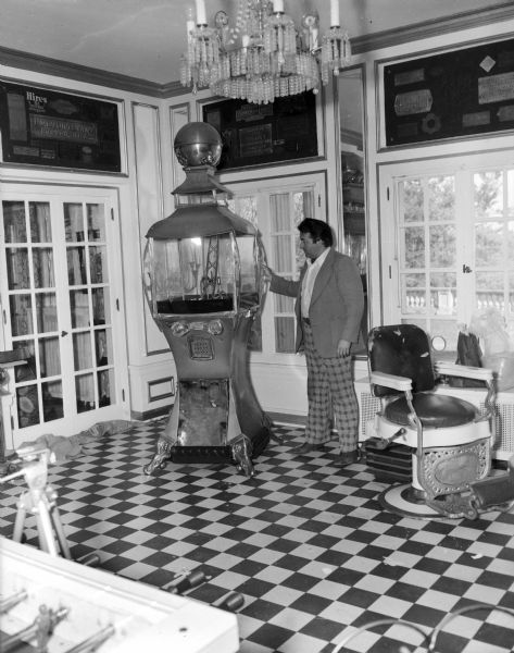 Tom Every (Doc Evermor) at his house in the Highlands. He is standing next to a large arcade claw crane game. The room housing has a checkered patterned floor, and metal name plates from old manufacturing companies are displayed on the walls above the windows. There is a barber chair on the right, and in the foreground on the left is the edge of a foosball table.