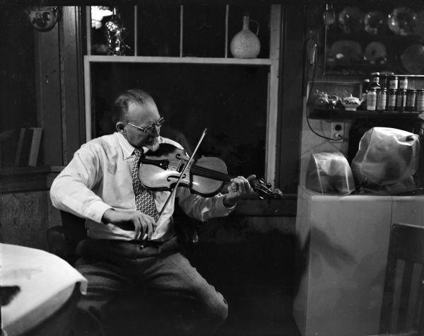 Man playing fiddle in the kitchen of a house.