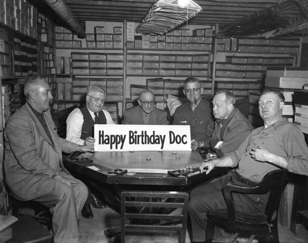 Group portrait of six men holding up a signboard saying "Happy Birthday Doc" in the middle of a card table. The group is sitting and playing cards in what appears to be a storage room, with boxes stacks on shelves around them.