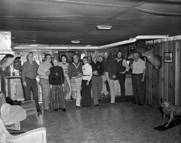 Group portrait of men and women posing together in front of a bar. The room has panelled walls, on which are mounted a number of deer heads and other paraphernalia. On the floor on the right is a taxidermied snarling animal on a wood base. Bob Bender is on the far right.