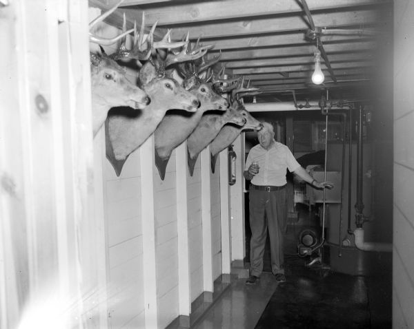 Portrait of Bob Bender holding a drink in his hand and standing near a wall of five mounted deer heads. He appears to be standing in a basement area.