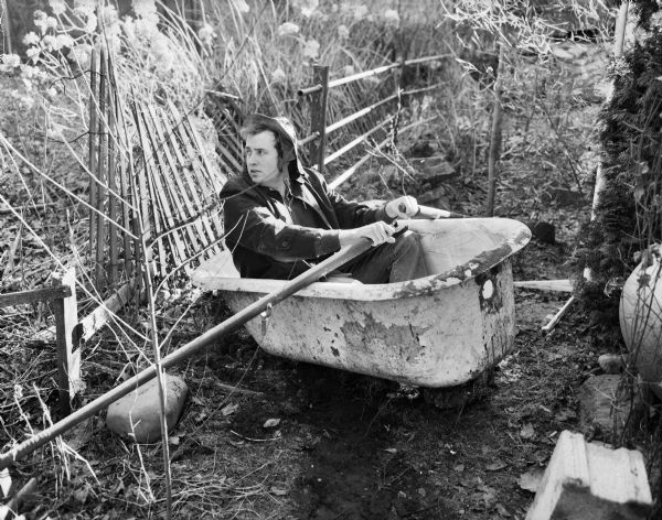 Curt Martin sitting in a clawfoot bathtub outdoors and holding oars as if the he was rowing. Behind the man are plants and a broken snow fence.