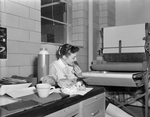 Bea Walusiak Heitmeyer is sitting and eating in the darkroom of the Gisholt Machine factory. On the table in the foreground is a thermos, coffee cup, wrapped food, and various papers.
