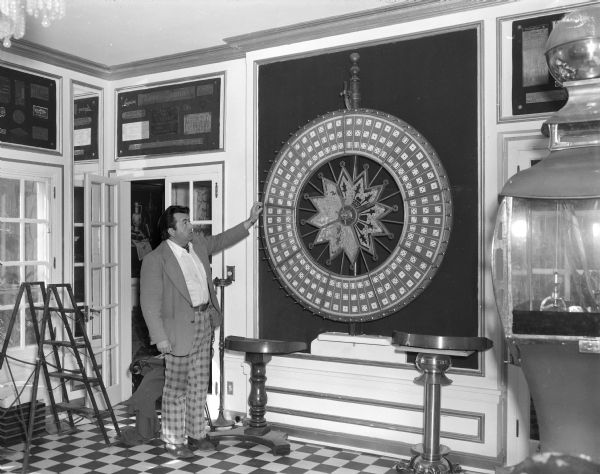 Tom Every (Doc Evermor) at his house in the Highlands. He is standing and holding one of the nickel plated pins on a wall-mounted Big Six Wheel or Wheel of Fortune with three dice in each segment. The center star of the wheel reads: "H.C. Evans & Co, Chicago." The star in the center is reverse painted glass. The room has a checkered patterned floor, and metal name plates from old manufacturing companies are displayed on the walls. On the far right is a large arcade claw crane game.