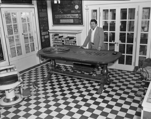 Tom Every (Doc Evermor) at his house in the Highlands. He is standing next to a roulette table. Behind him are french doors, and on the wall behind him are boxes containing rolls of player piano music lining the shelves of a bookcase. On the far left is a barber chair, and in the right foreground is part of a foosball table. The room has a checkered patterned floor, and metal name plates from old manufacturing companies are displayed on the walls.