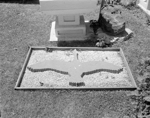 Prone flat sculpture of a goose in flight set in a bed of white gravel in the lawn of Sid's backyard. It is a gray slab cut to look like a flying bird. The white square base of the "Three-Tiered Fountain" and two stacked decorative rocks are in the background.