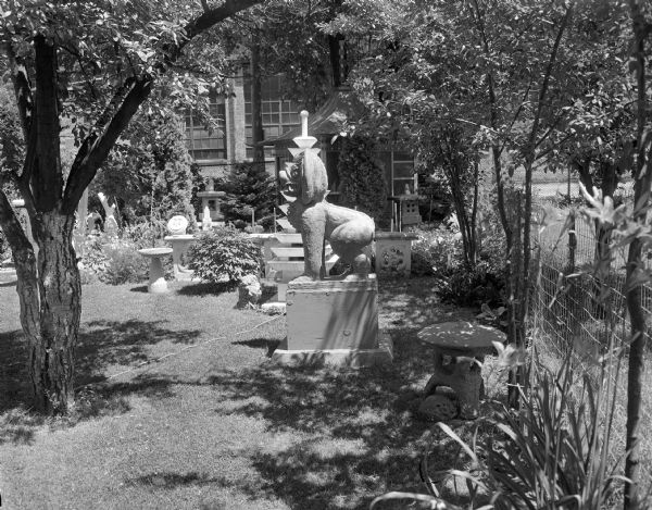 View looking into Sid's backyard with his concrete sculptures set among the trees and plants bordering the lawn. A large dog-lion sculpture is in the foreground. There is a wire fence on the right, and the Madison-Kipp factory building is in the background. 
