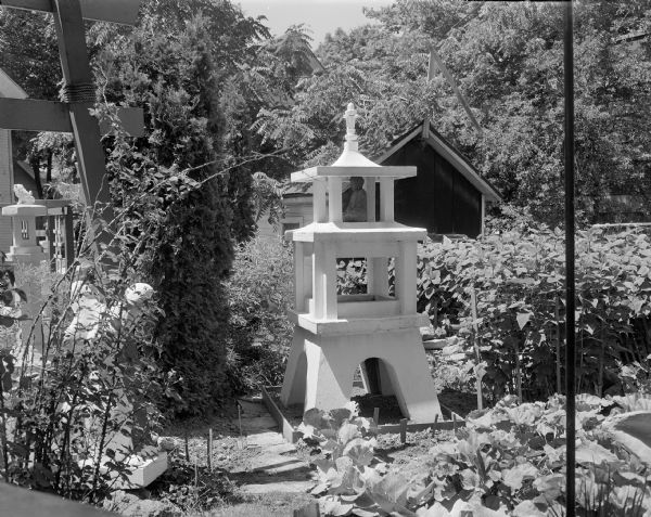 View across Sid's backyard with a shed structure in the background. In the yard are plants, trees, and sculptures which are surrounding a large, white, lantern-like structure, which has a Buddha sculpture inside. The Madison-Kipp Corporation factory building, obscured by trees, is in the far background. 