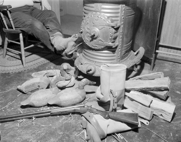Sid sitting on the left in a chair, with only his legs and feet visible, resting his feet on a woodburning stove. In the foreground is a pile of wooden duck decoys and firewood, along with a pair of waders and a rifle.