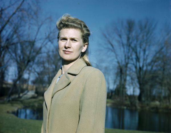 Waist-up portrait of Bernice Boyum Bergner, Sid Boyum's sister, wearing a beige coat. In the background are trees and a pond.