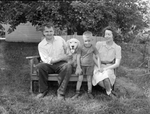 Group portrait of Sid, his wife Margaret, son Steve, and dog Duke, posing outdoors on a bench, perhaps in Sid's backyard. Sid, wearing a collared shirt, is sitting on the left side of the bench, with his dog in the center. Margaret, wearing a striped dress, is sitting on the bench on the right (eyes closed). Between them their son Steve is standing, who is wearing a t-shirt and shorts.